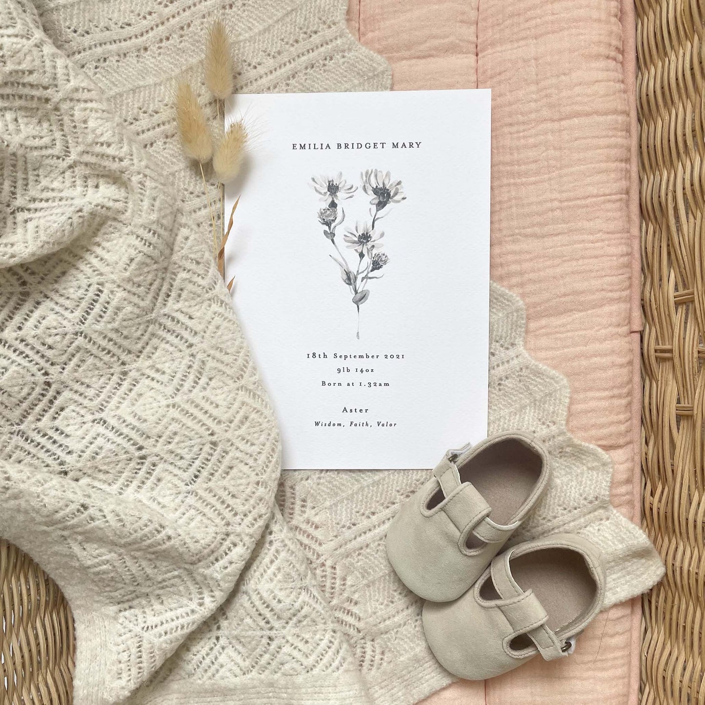 close up lifestyle shot of personalised birth flower baby print featuring a watercolour print of an aster for septembers birth month. laid in a changing basket with baby shoes, blanket and bunny tail dried flowers.