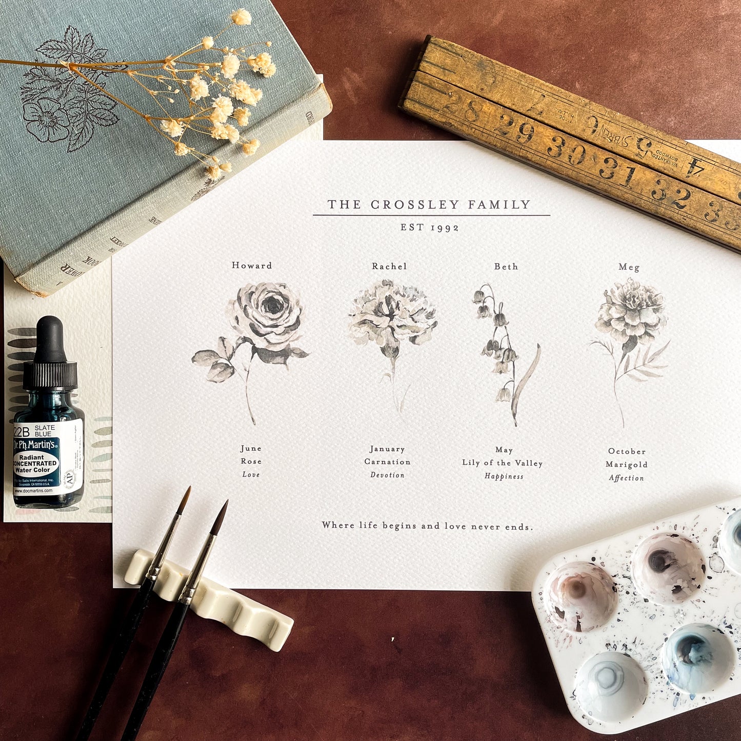 A flat lay image showing Kester Studio's personalised birth flower family print with four birth flowers. Surround by painting supplies, a wooden ruler and dried gyposphila.