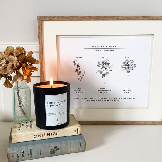 Lifestyle image showing personalised birth flower grandchildren print in an oak frame with double mount. 3 birth flowers feature on the print with their symbolic meanings shown. Lily of the Valley for Happiness, Chrysanthemum for Cheerfulness and Cranation for Devotion. Frame is surrounded by books and a lit candle.